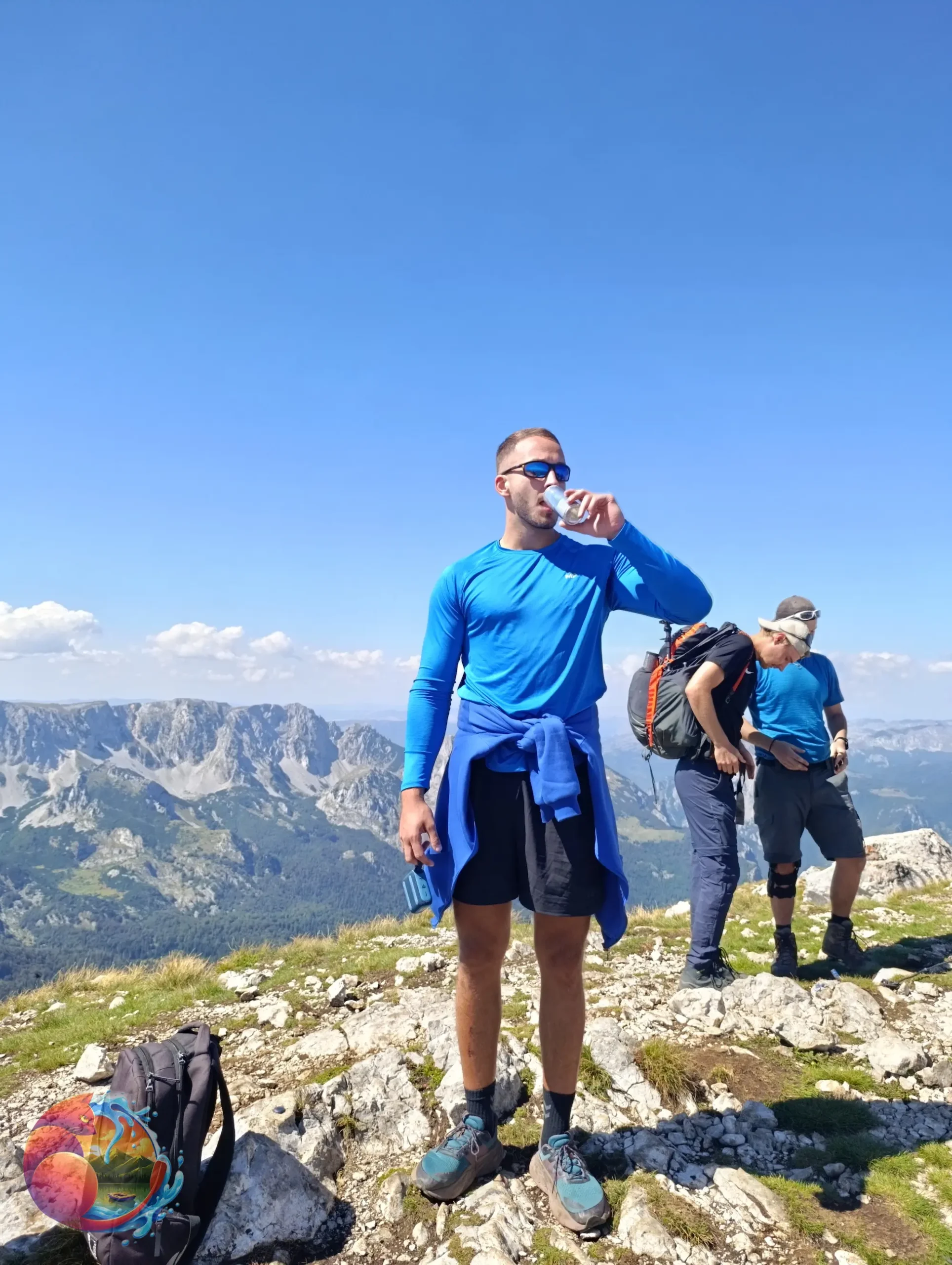 Naslovna A man stands on a mountain, wearing a backpack and holding a bottle of water, enjoying the scenic view.