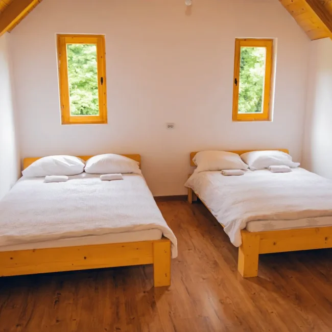 A cozy bedroom featuring two wooden beds with white linens and pillows, illuminated by natural light from two small windows.