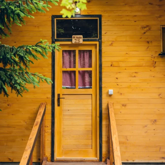 Front view of a rustic wooden cabin with a sloped roof, featuring four windows and a wooden door, surrounded by trees.
