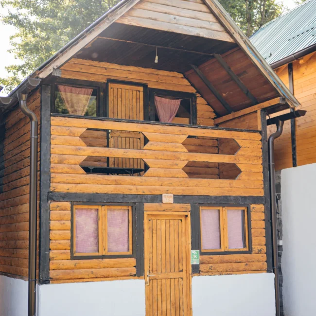 A wooden cabin with a sloped roof, featuring a front door, multiple windows, and decorative wooden elements.