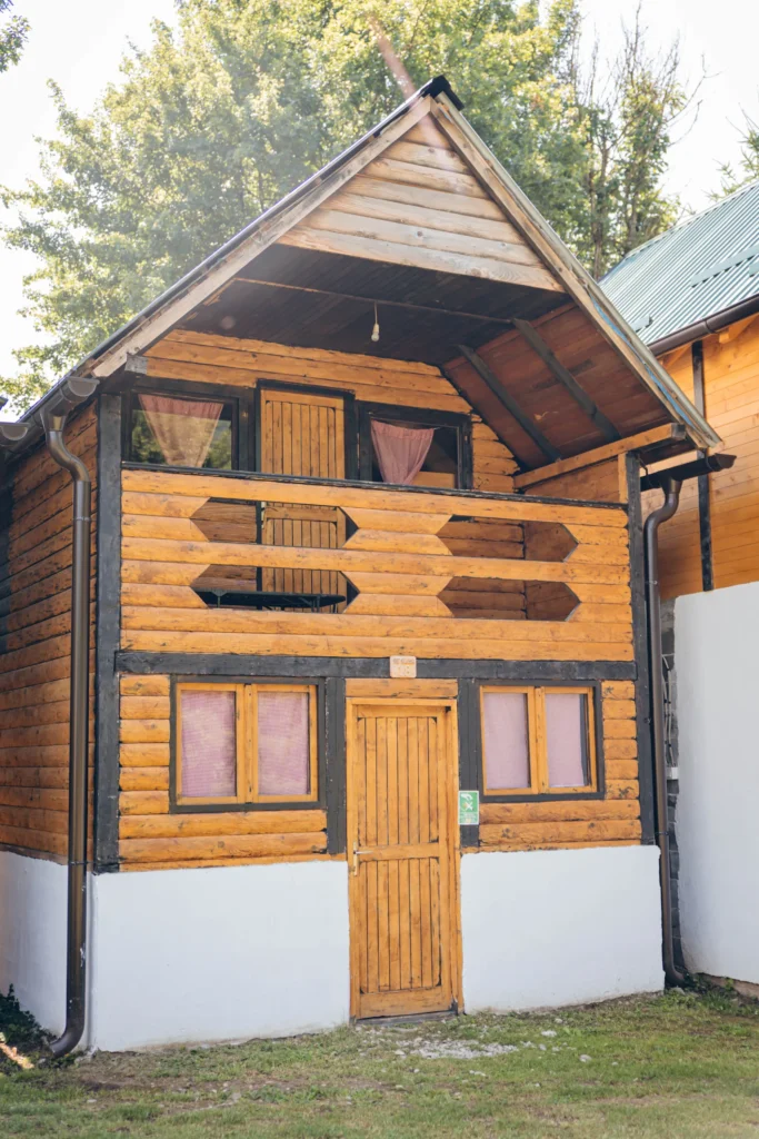 A wooden cabin with a sloped roof, featuring a front door, multiple windows, and decorative wooden elements.